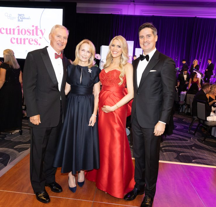 Photograph of four people in black tie gala attire for the 2023 Children's Ball: Curiosity Cures; Left to right: J. Christopher Reyes (Medical Center Board) wearing a full black tuxedo, Anne Reyes wearing a black ankle length flair midi dress, and Event Co-chairs Elizabeth wearing a red strapless gown cradling what looks like a baby bump and Andrew Reyes wearing a full tuxedo with bow tie;