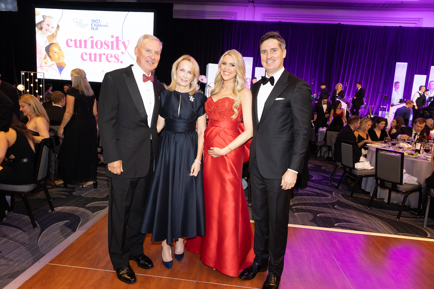 Photograph of four people in black tie gala attire for the 2023 Children's Ball: Curiosity Cures; Left to right: J. Christopher Reyes (Medical Center Board) wearing a full black tuxedo, Anne Reyes wearing a black ankle length flair midi dress, and Event Co-chairs Elizabeth wearing a red strapless gown cradling what looks like a baby bump and Andrew Reyes wearing a full tuxedo with bow tie;